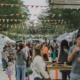 A busy outdoor market with people walking, eating, and shopping among white tents. Colorful bunting hangs above. Trees line the walkway, and families, couples, and vendors fill the lively scene.